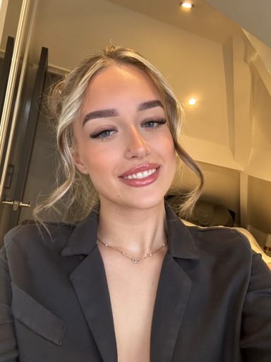Smiling young woman with blonde hair, wearing a grey blazer, in a well-lit room.